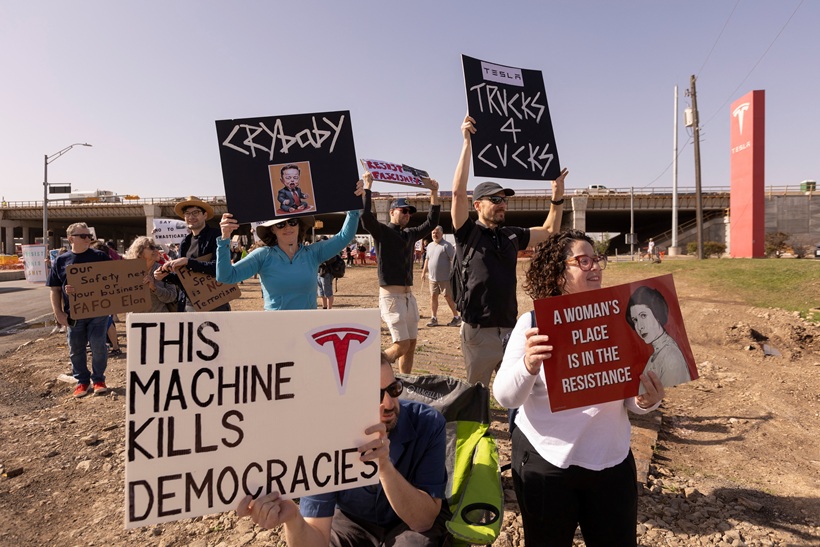 Pessoas em frente a uma fábrica da Tesla carregam cartazes com textos em inglês. No destaque, um cartaz em que se lê, em tradição livre: "Esta máquina mata democracias", com um logo da Tesla ao lado